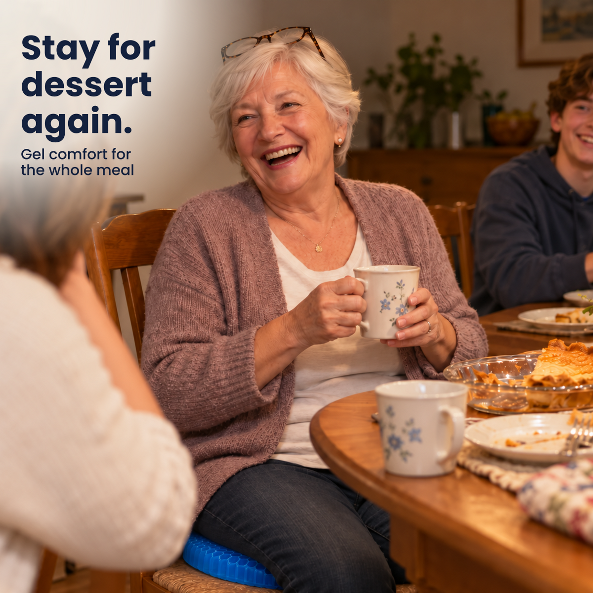 Smiling grandmother at the family dinner table with a cup of tea, sitting on the blue honeycomb gel cushion. Text: Stay for dessert again.